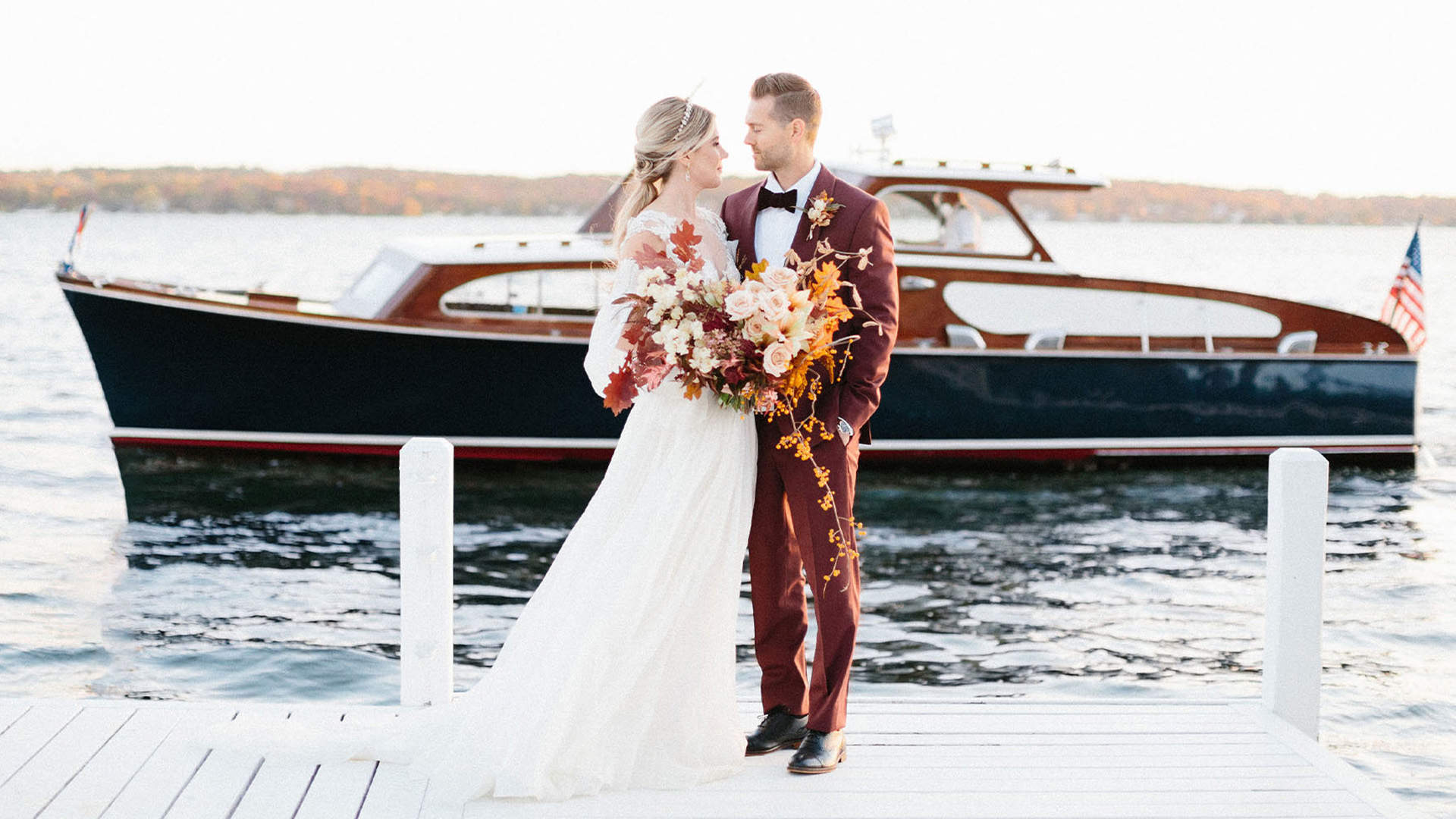 Couple embracing on a white dock before a navy vintage boat on Lake Geneva, bride holding an autumn bouquet